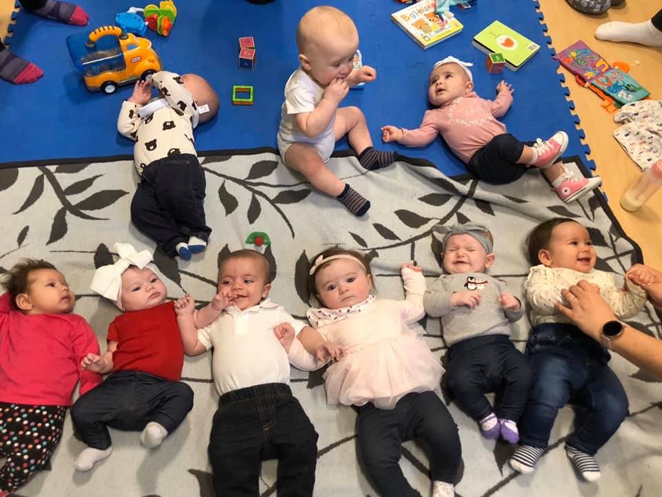 Seven babies lying side by side on a play mat