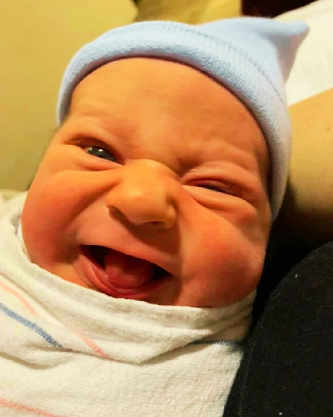 A newborn in a hospital blanket and cap, grinning