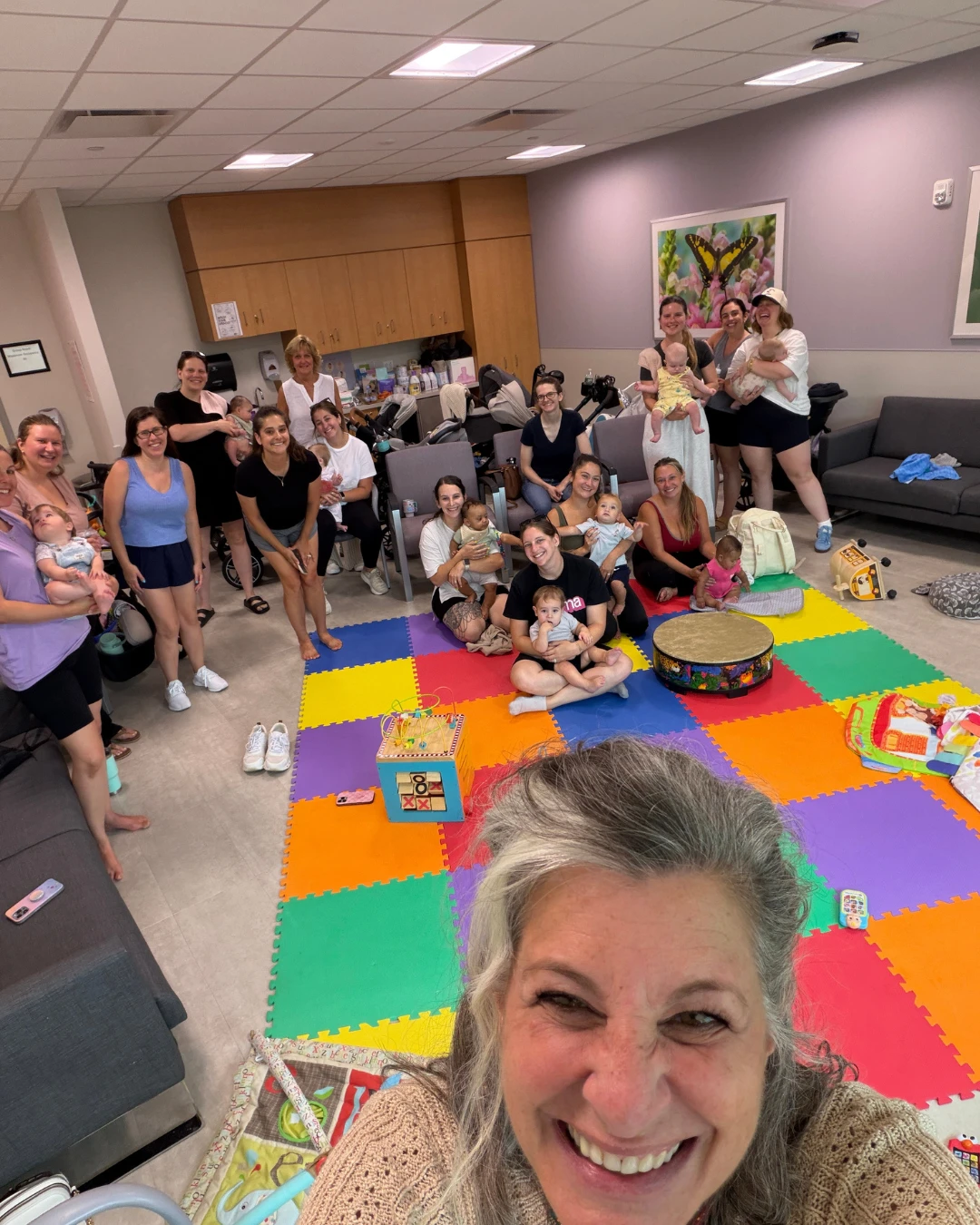 A room full of smiling moms and babies gathered around a colorful play mat